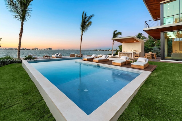 pool at dusk with outdoor lounge area, a water view, and a yard