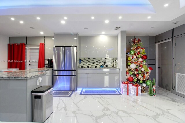 kitchen featuring sink, decorative backsplash, gray cabinets, light stone countertops, and stainless steel refrigerator