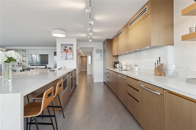 kitchen with rail lighting, light stone counters, stainless steel oven, a breakfast bar, and sink