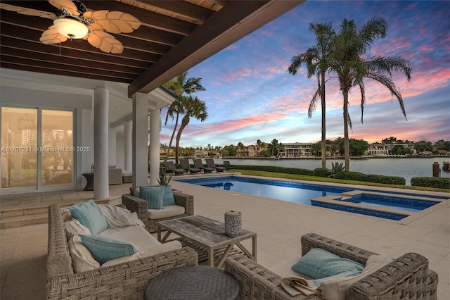 pool at dusk with an in ground hot tub, a water view, ceiling fan, and a patio