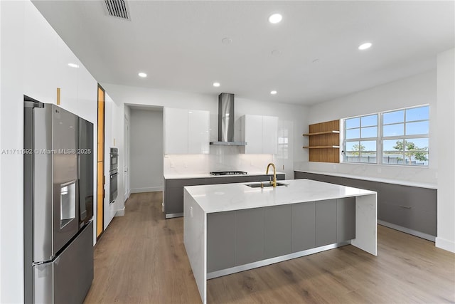 kitchen featuring sink, a kitchen island with sink, black appliances, white cabinets, and wall chimney exhaust hood