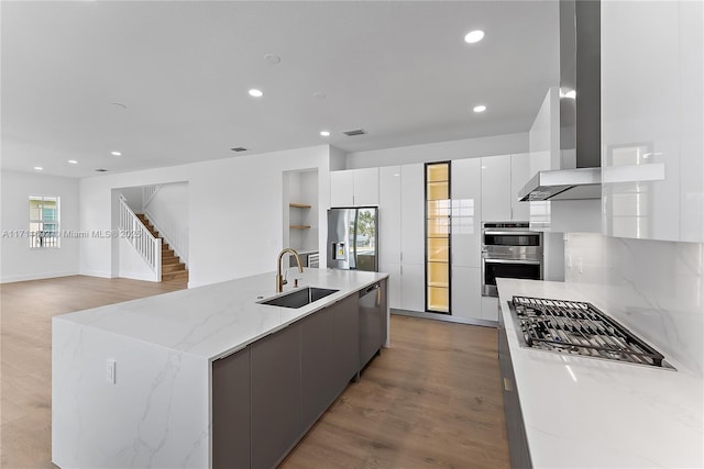 kitchen with white cabinetry, an island with sink, sink, stainless steel appliances, and wall chimney exhaust hood