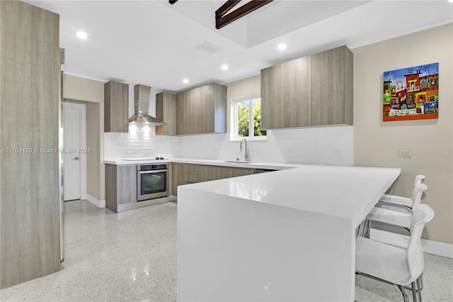 kitchen with kitchen peninsula, tasteful backsplash, wall chimney exhaust hood, stainless steel oven, and a breakfast bar