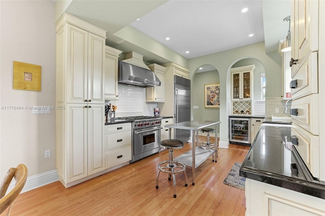 kitchen featuring high quality appliances, beverage cooler, light wood-style floors, wall chimney range hood, and baseboards