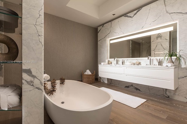 full bathroom with double vanity, wood finished floors, a freestanding bath, stone wall, and a sink