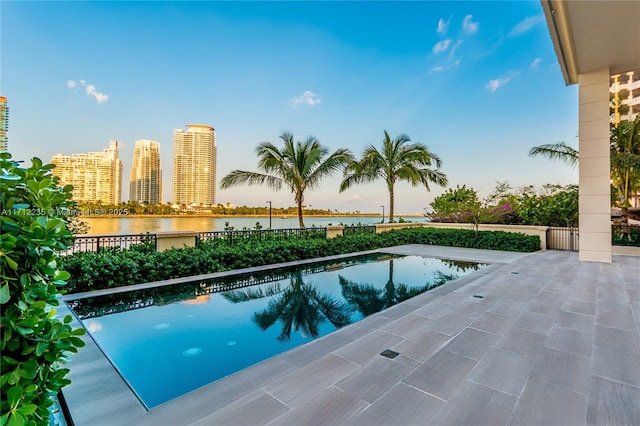 pool at dusk featuring a patio area, a water view, a fenced in pool, and a city view