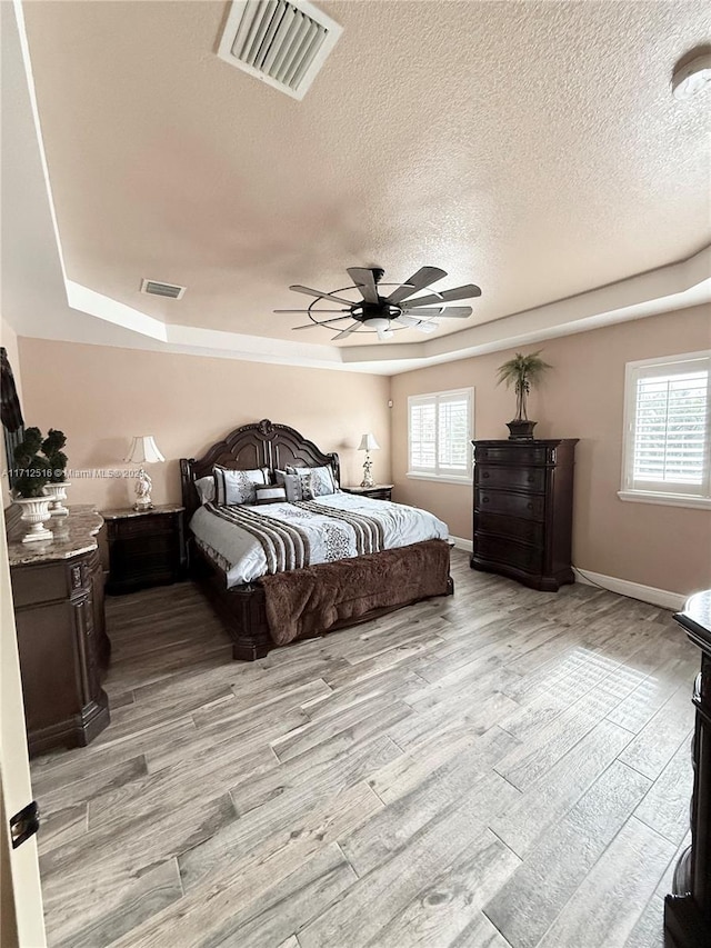 bedroom with a textured ceiling, ceiling fan, light hardwood / wood-style flooring, and a tray ceiling