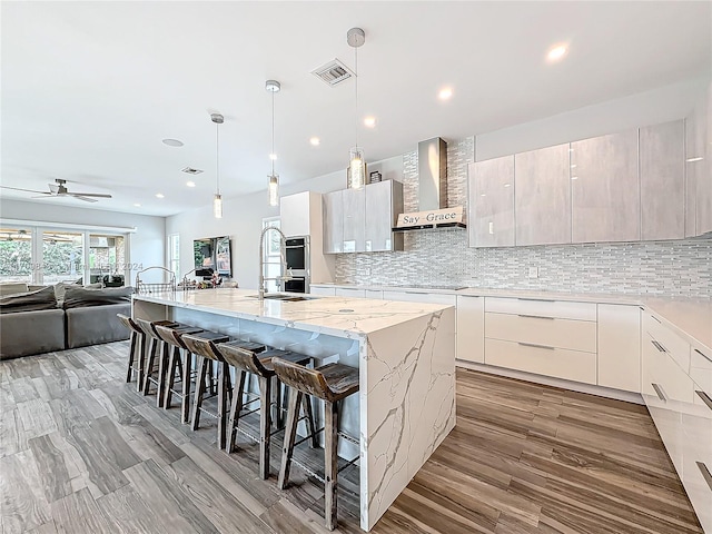 kitchen with ceiling fan, wall chimney exhaust hood, a kitchen breakfast bar, an island with sink, and decorative light fixtures