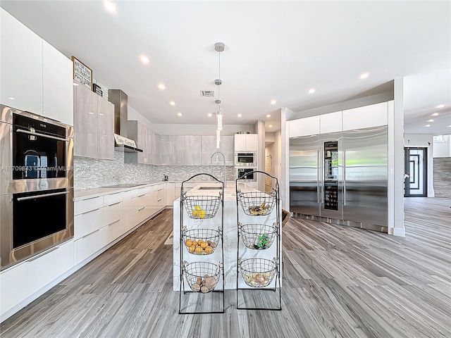 kitchen featuring appliances with stainless steel finishes, light wood-type flooring, wall chimney exhaust hood, white cabinets, and hanging light fixtures