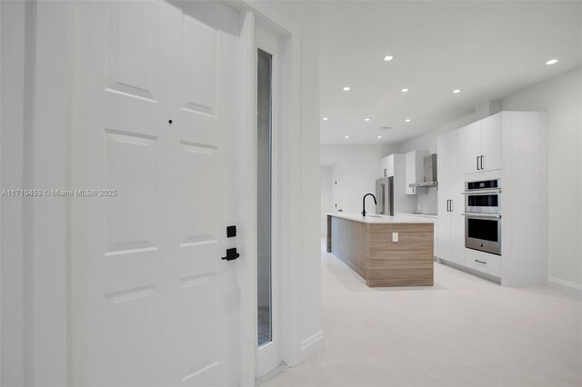 kitchen featuring white cabinetry, sink, stainless steel appliances, wall chimney range hood, and a kitchen island with sink