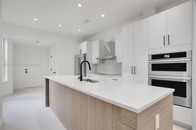 kitchen featuring sink, wall chimney exhaust hood, a kitchen island with sink, white cabinets, and appliances with stainless steel finishes