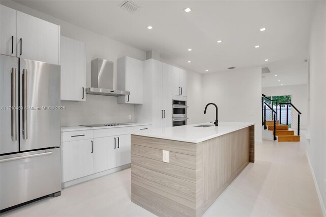 kitchen featuring stainless steel appliances, wall chimney range hood, white cabinets, a kitchen island with sink, and light tile patterned floors