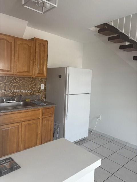 kitchen featuring light tile patterned flooring, a sink, decorative backsplash, freestanding refrigerator, and brown cabinets