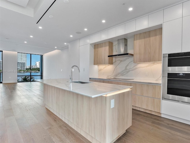 kitchen with sink, wall chimney range hood, a large island with sink, light hardwood / wood-style flooring, and white cabinets