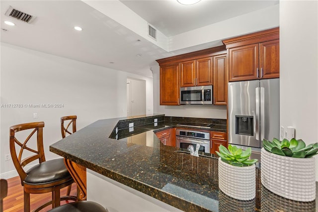 kitchen with stainless steel appliances, kitchen peninsula, dark stone countertops, wood-type flooring, and a breakfast bar