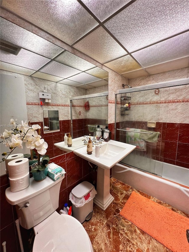 bathroom with a paneled ceiling, toilet, shower / bath combination with glass door, and tile walls