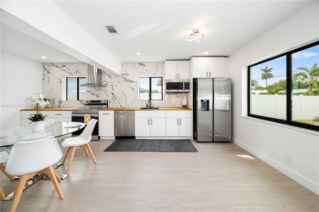 kitchen featuring decorative backsplash, stainless steel appliances, wall chimney range hood, white cabinets, and light hardwood / wood-style floors