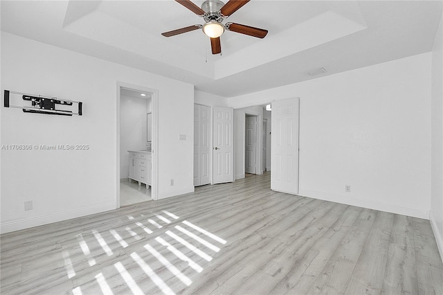 unfurnished bedroom featuring ceiling fan, light hardwood / wood-style floors, a tray ceiling, and ensuite bath