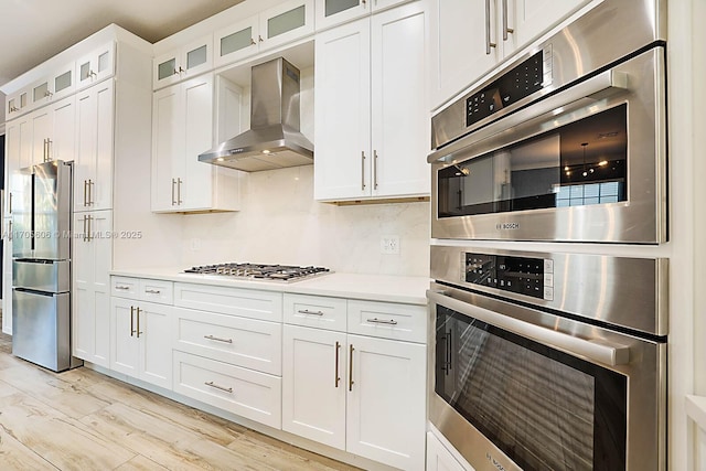 kitchen with wall chimney range hood, light hardwood / wood-style flooring, appliances with stainless steel finishes, tasteful backsplash, and white cabinetry