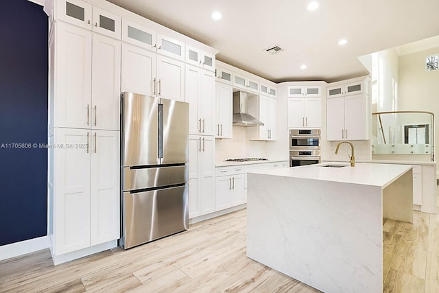 kitchen with appliances with stainless steel finishes, white cabinetry, a kitchen island with sink, and wall chimney exhaust hood