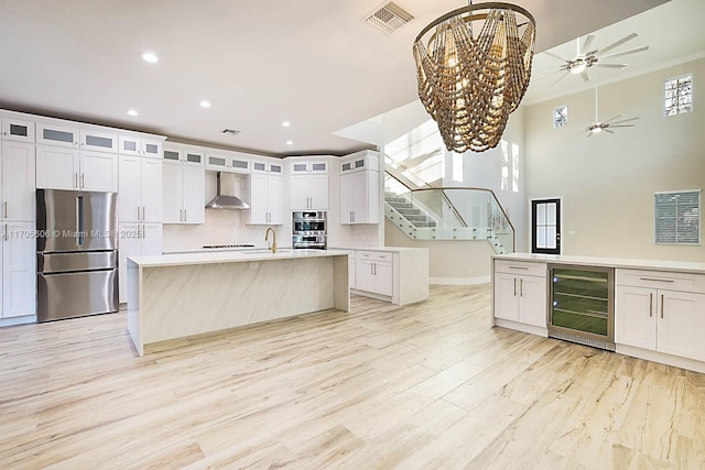 kitchen with white cabinets, wall chimney exhaust hood, an island with sink, appliances with stainless steel finishes, and beverage cooler