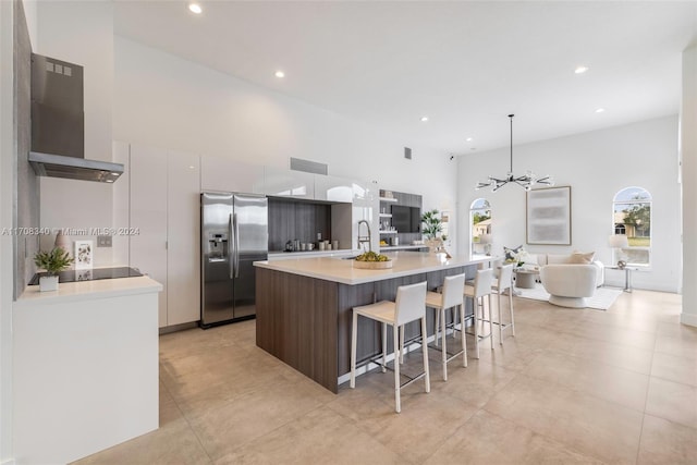 kitchen featuring dark brown cabinetry, a center island with sink, decorative light fixtures, stainless steel fridge with ice dispenser, and range hood