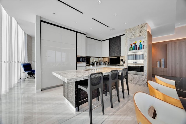 kitchen featuring a kitchen island with sink, a kitchen breakfast bar, sink, light stone counters, and white cabinetry