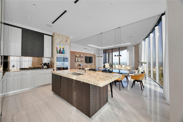 kitchen featuring white cabinetry, sink, light stone countertops, expansive windows, and an island with sink