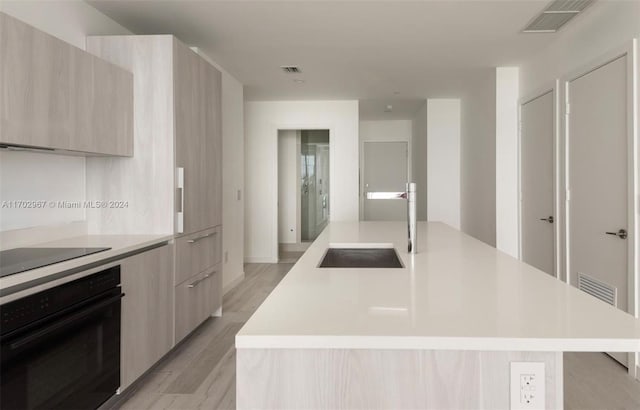 kitchen featuring light brown cabinetry, a center island with sink, black appliances, and light hardwood / wood-style floors