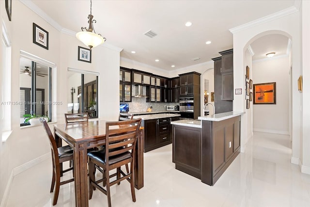 kitchen with dark brown cabinets, wall chimney range hood, decorative light fixtures, decorative backsplash, and kitchen peninsula