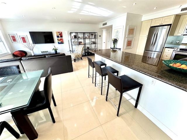 kitchen featuring dark stone counters, crown molding, appliances with stainless steel finishes, light tile patterned flooring, and a breakfast bar area