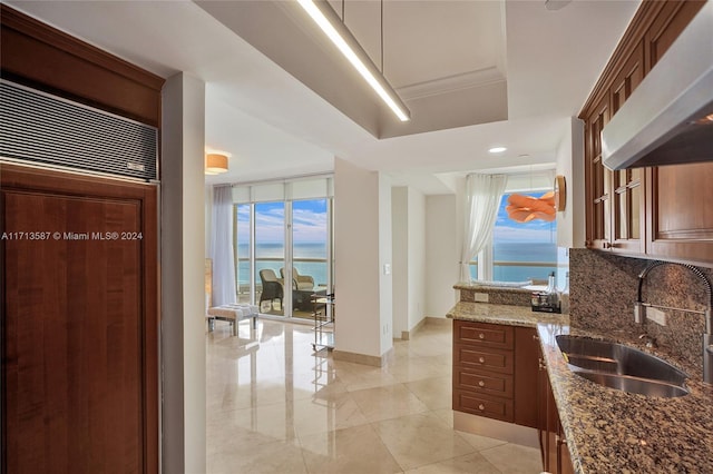 kitchen with wall chimney exhaust hood, dark stone countertops, sink, backsplash, and a water view