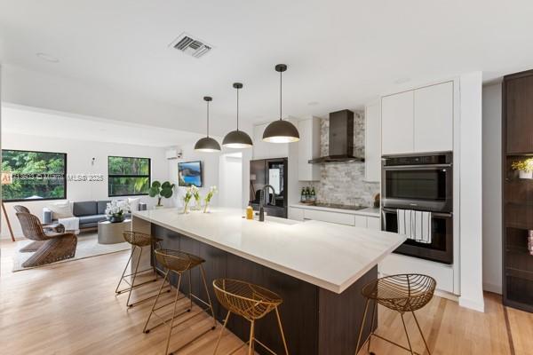 kitchen with white cabinetry, wall chimney exhaust hood, a kitchen breakfast bar, and a center island with sink