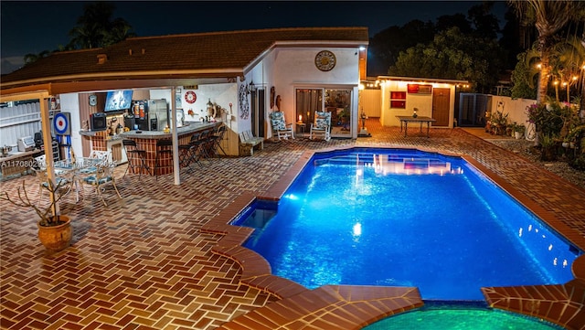 pool at night with a patio area, a pool with connected hot tub, outdoor dry bar, and fence