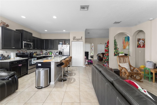 kitchen with a kitchen bar, appliances with stainless steel finishes, light stone counters, a kitchen island with sink, and light tile patterned floors