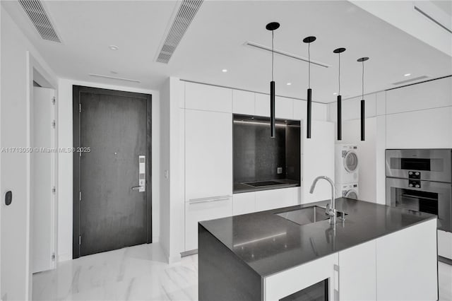 kitchen with a kitchen island with sink, sink, stacked washer and dryer, white cabinetry, and hanging light fixtures