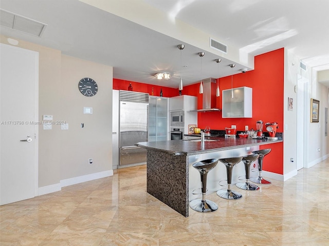 kitchen featuring a kitchen bar, kitchen peninsula, wall chimney range hood, built in appliances, and hanging light fixtures