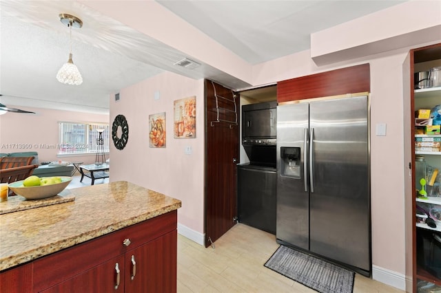 kitchen featuring light stone countertops, stainless steel refrigerator with ice dispenser, ceiling fan, stacked washer / dryer, and hanging light fixtures