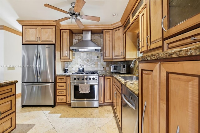 kitchen featuring wall chimney range hood, ceiling fan, dark stone countertops, appliances with stainless steel finishes, and tasteful backsplash