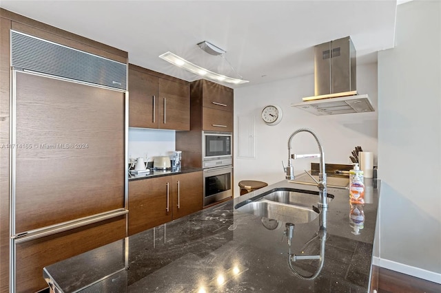 kitchen featuring built in appliances, sink, dark stone counters, and wall chimney range hood