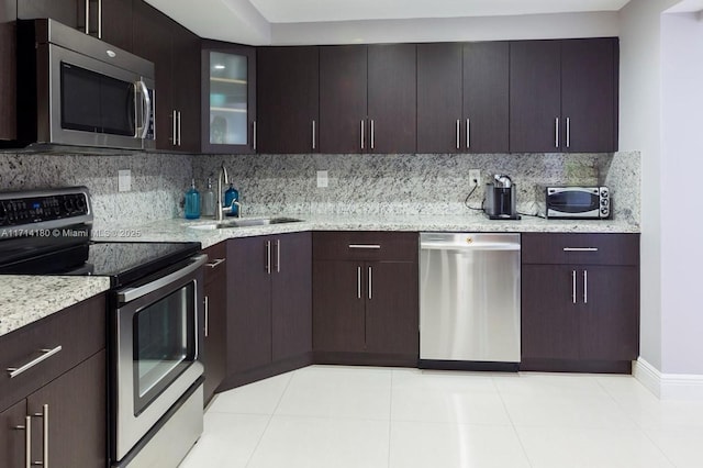 kitchen with stainless steel appliances, a sink, dark brown cabinets, decorative backsplash, and glass insert cabinets