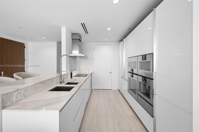 kitchen with white cabinets, black electric stovetop, wall chimney range hood, sink, and light stone counters