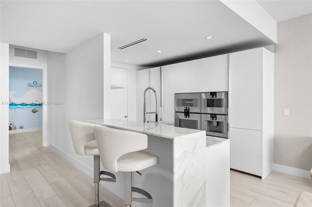 kitchen featuring light stone countertops, double oven, light hardwood / wood-style flooring, white cabinetry, and a breakfast bar area