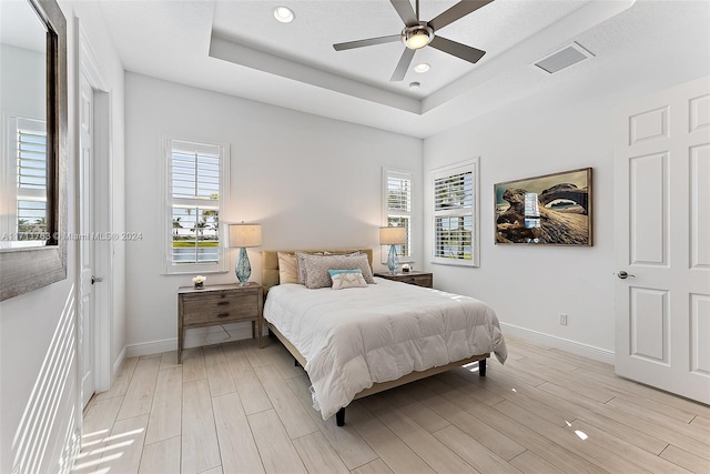 bedroom with multiple windows, light hardwood / wood-style floors, a raised ceiling, and ceiling fan