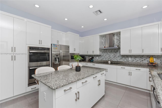 kitchen with stainless steel appliances, a kitchen island, wall chimney range hood, light tile patterned floors, and white cabinetry