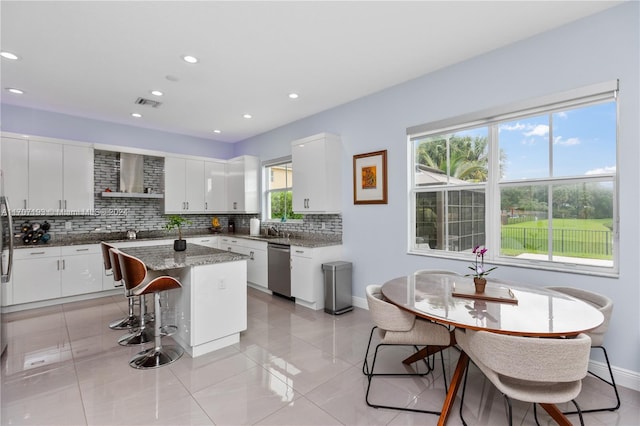 kitchen featuring dishwasher, wall chimney exhaust hood, a kitchen island, dark stone countertops, and white cabinets