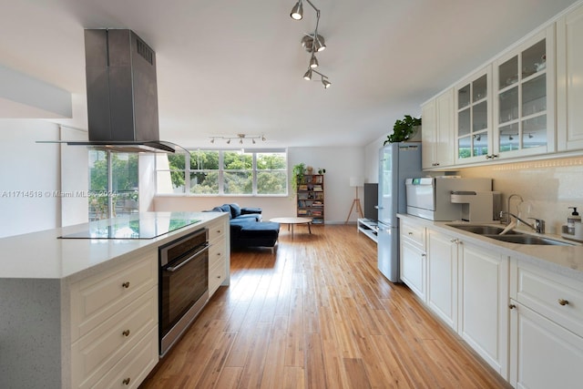 kitchen featuring stainless steel oven, white cabinets, sink, light wood-type flooring, and island range hood