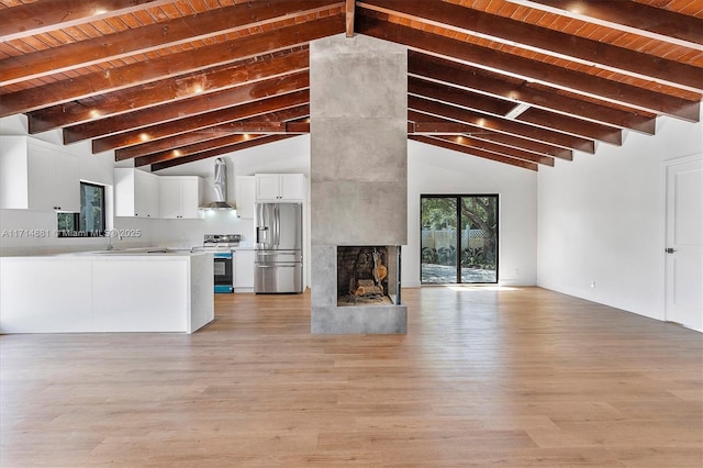 kitchen featuring a multi sided fireplace, appliances with stainless steel finishes, white cabinetry, vaulted ceiling with beams, and wall chimney range hood