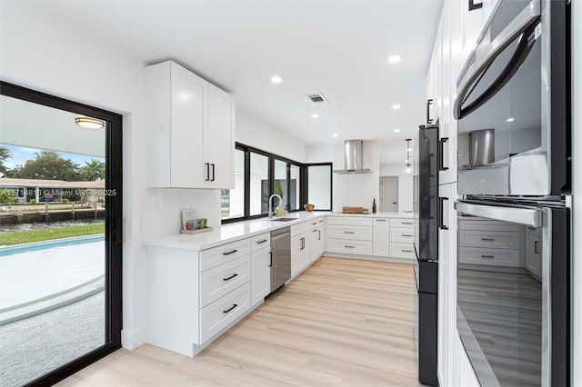 kitchen with sink, wall chimney range hood, light hardwood / wood-style flooring, stainless steel dishwasher, and white cabinets