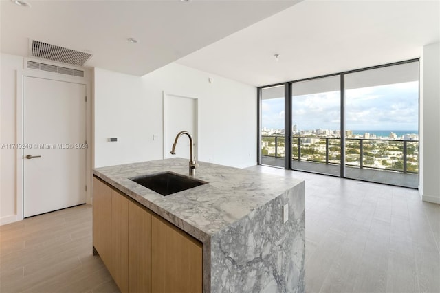 kitchen featuring floor to ceiling windows, sink, light wood-type flooring, an island with sink, and light stone counters